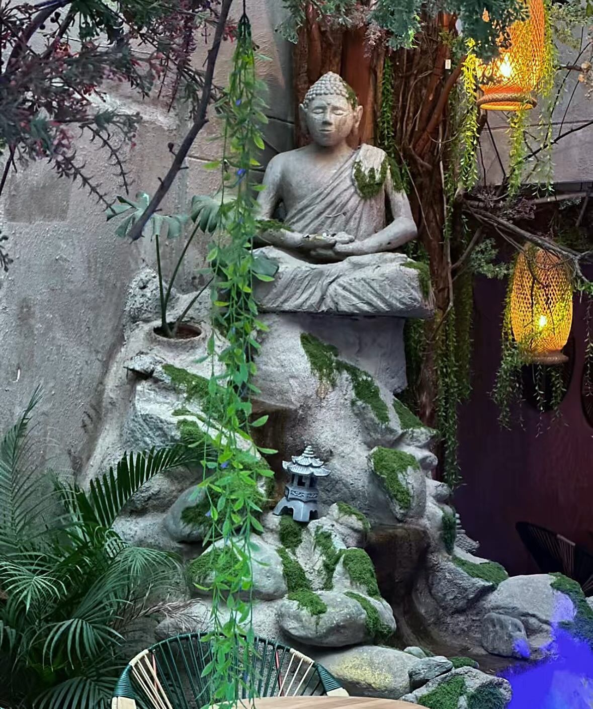 Buda de piedra sobre una cascada entre plantas y linternas, en la llegada a Mystic